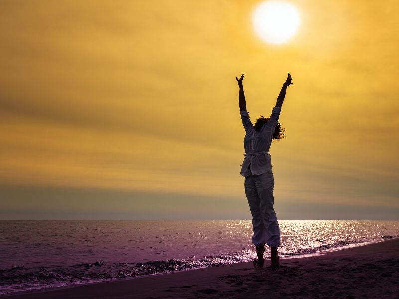 Eine Frau am Strand bei Sonnenuntergang streckt sich vor Freude. Barbara Erdt kümmert sich um ganzheitliche Gesundheit in Mengkofen.